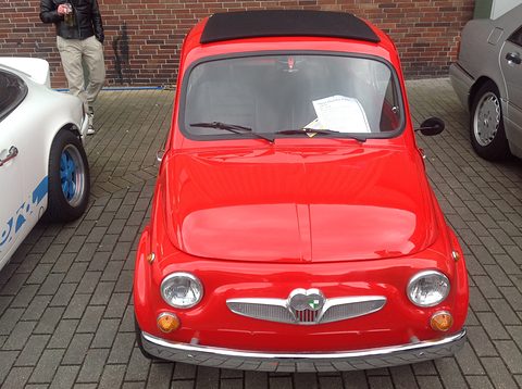Red small two-door car from the front, black fabric roof closed, parked on brick paving between a white sports car and a silver saloon. Opens a larger view