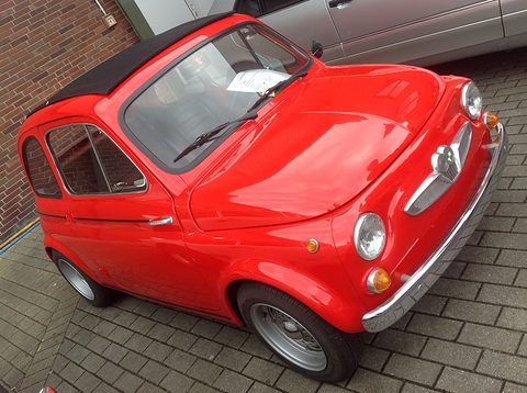 High view of a glossy red small car’s bonnet, windscreen and nose, with chrome grille, round headlamps and orange indicators. Opens a larger view