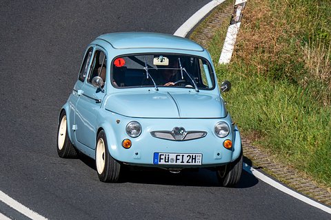 Light blue small car with cream wheels and a red number 1 sticker on the windscreen, cornering beside long grass. Opens a larger view