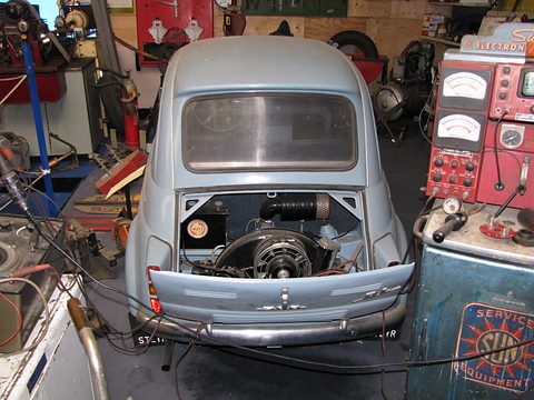 Pale blue small car seen from behind with the engine lid open, showing a black fan housing and air filter, cables draped over the tail. Opens a larger view
