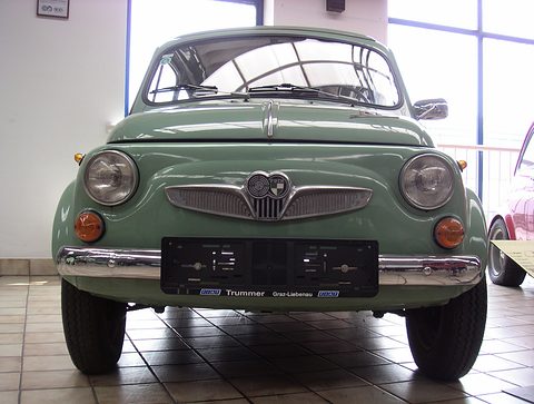 Pale green small car head-on on a tiled floor, round headlamps, winged chrome grille with twin badge, empty number-plate frame. Opens a larger view