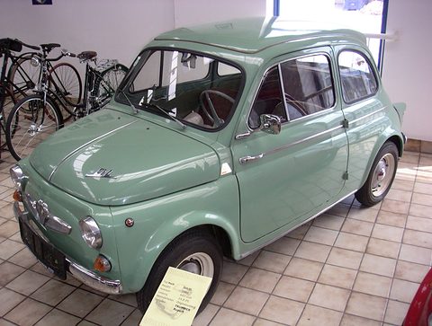 Pale green small saloon with chrome trim and ‘DL’ script on the bonnet, on a tiled floor beside vintage bicycles, museum card in front.