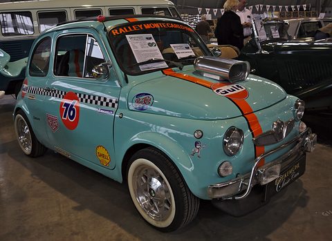 Pale blue small saloon with orange stripe, Gulf, 76 and Shell decals, bonnet scoop and nudge bar, at an indoor show. Opens a larger view
