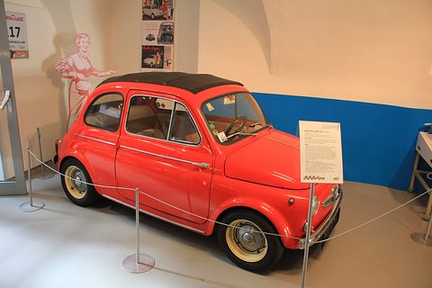 Red small saloon with black fabric roof and cream wheels behind a rope barrier, information board beside it, period posters on the wall. Opens a larger view