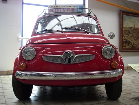 Red small car seen head-on on tiles, winged grille with twin badge, roof rack with an L-plate driving-school sign. Opens a larger view