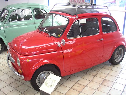 Red small saloon with chrome roof rack carrying a driving-school sign, on a tiled museum floor beside a pale green car. Opens a larger view