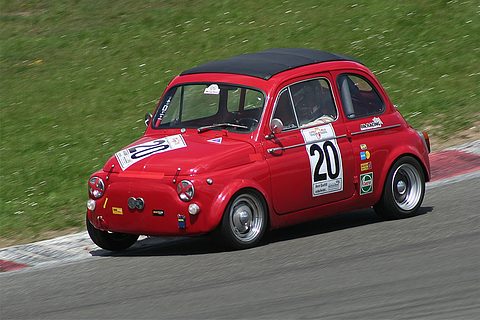 Red small saloon with race number 20 and sponsor decals cornering on a kerbed circuit beside a grass bank. Opens a larger view