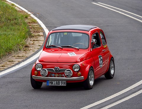 Red small car with black fabric roof, two large round lamps and badges along the front bumper, on a curving road. Opens a larger view
