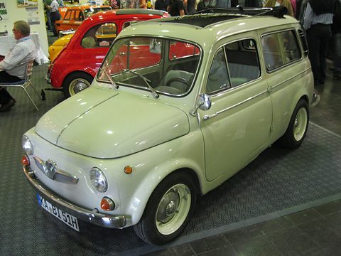 Pale cream small estate car with a rolled-back fabric roof and winged chrome grille, on a show-hall floor beside a red saloon. Opens a larger view