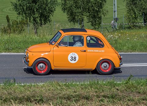 Orange small car with red wheels and race number 38 driving past a grassy verge, helmeted driver at the wheel. Opens a larger view