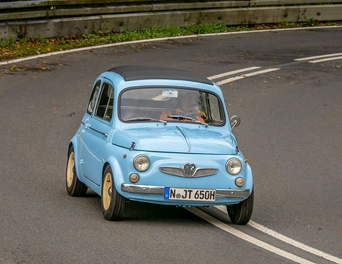 Light blue small car with a winged chrome grille and cream wheels, driver in an orange top, rounding a bend. Opens a larger view