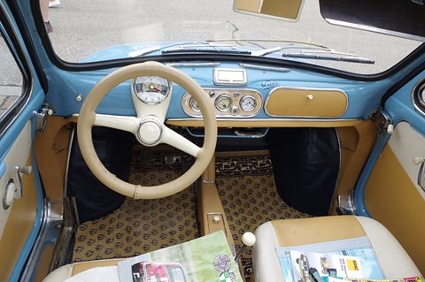 Car interior seen through the open door: blue dashboard, cream steering wheel, patterned floor mats and leaflets on the seats. Opens a larger view