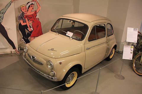 Cream small saloon with yellow wheels and ‘D’ script on the bonnet, roped off in a museum beside cartoon dancer cut-outs. Opens a larger view