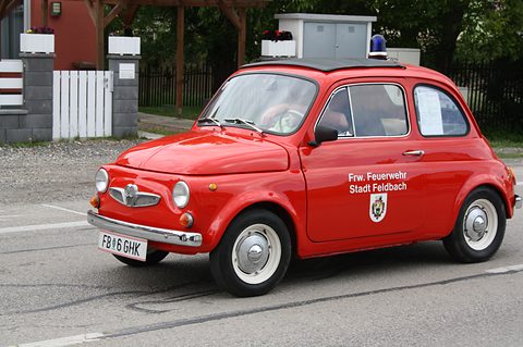 Red small saloon with blue roof beacon and ‘Frw. Feuerwehr Stadt Feldbach’ lettering and crest on the door, on a village road. Opens a larger view