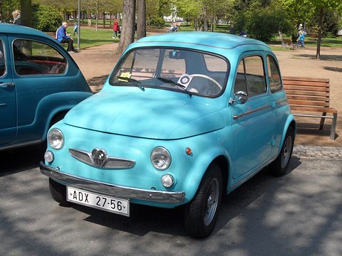 Turquoise small saloon with winged grille and Czech plate in dappled shade, park benches and trees behind. Opens a larger view