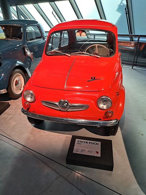 Red small car from the front on a grey museum floor, winged grille, ‘D’ script on the bonnet, information block in front. Opens a larger view