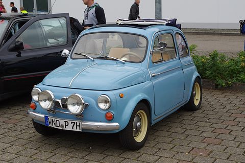 Light blue small car with cream wheels and three round lamps on the front bumper, parked on block paving beside a black car. Opens a larger view