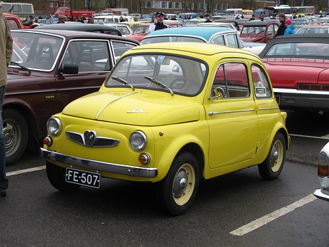 Yellow small saloon with cream wheels and ‘DL’ script on the bonnet, among other classic cars in a wet car park. Opens a larger view