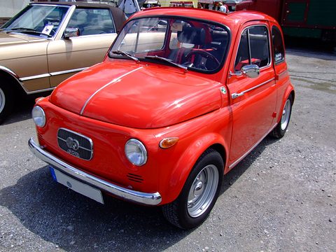 Bright red small rear-engined saloon on gravel, chrome bumper, oval grille with twin badge, parked beside a gold convertible.