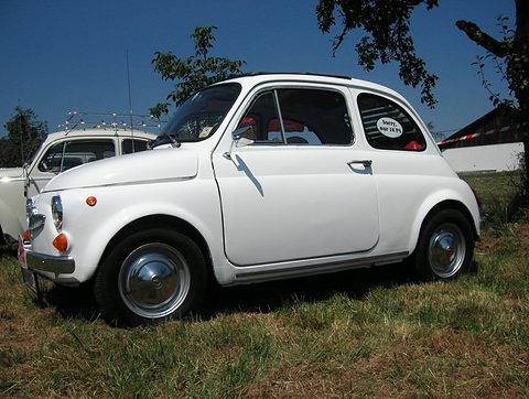 White small saloon parked on grass under a deep blue sky, folded fabric roof, tree branches above. Opens a larger view