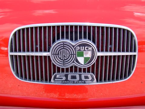 Close view of an oval chrome grille with vertical bars on a red car, carrying a linked Steyr and Puch badge and a 500 plate. Opens a larger view