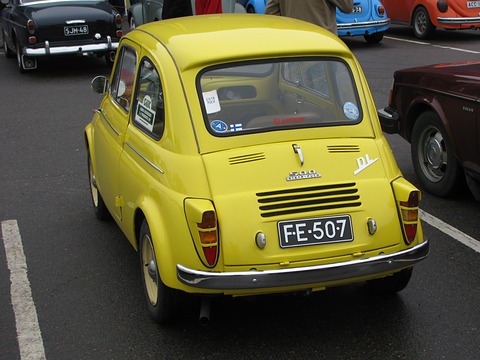 Rear of a yellow small saloon with louvred engine lid, chrome bumper and Finnish number plate, on a wet tarmac car park. Opens a larger view