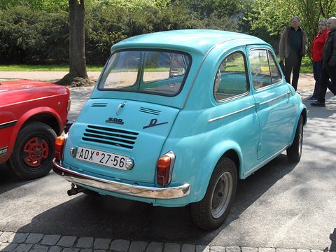 Rear three-quarter of a turquoise small saloon with louvred engine lid, chrome bumper and exhaust, in a sunny park. Opens a larger view