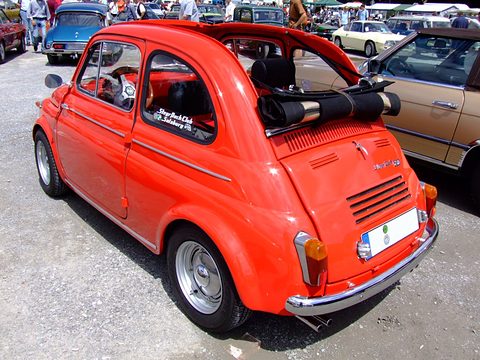 Red small saloon seen from the rear quarter, roof rolled back, twin exhaust pipes, Steyr-Puch script on the engine lid, show field behind. Opens a larger view
