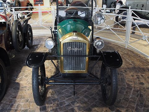 Front view of a very narrow green car with a brass radiator lettered Peugeot, two lamps and black wings on cobbles.