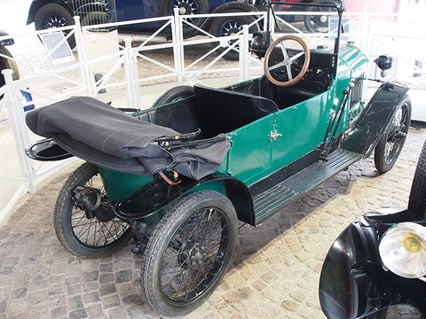A narrow green open car with a black folded hood on its tail, a long wooden-rimmed steering wheel and tandem seats.