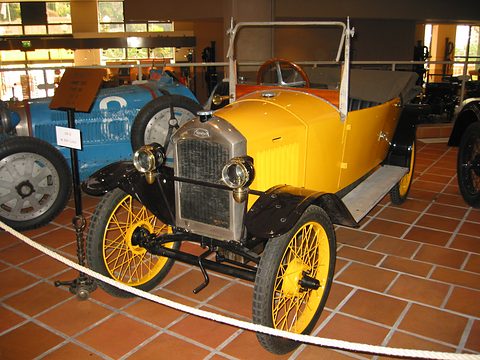 A narrow yellow open car with a nickel radiator, black wings and yellow wire wheels on red tiles behind a rope. Opens a larger view