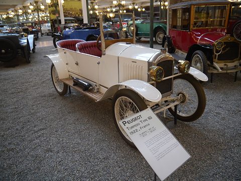 A white open two-seater with red seats and wire wheels on gravel, a printed placard leaning in front. Opens a larger view