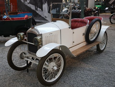 A white open two-seater with a black radiator, red buttoned seats and white wire wheels on grey gravel. Opens a larger view