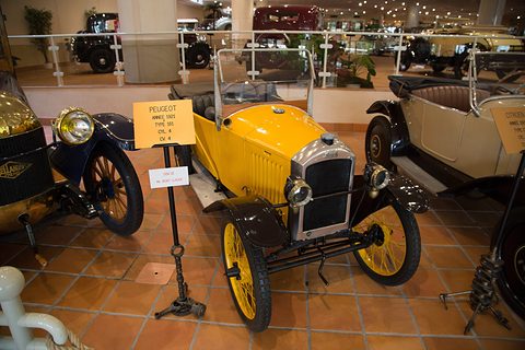A yellow narrow open car with black wings and yellow wire wheels beside a placard on a stand, on terracotta tiles. Opens a larger view