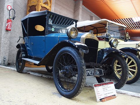 A blue two-seater with black wings, a white hood and side curtains, a placard on the floor before it. Opens a larger view