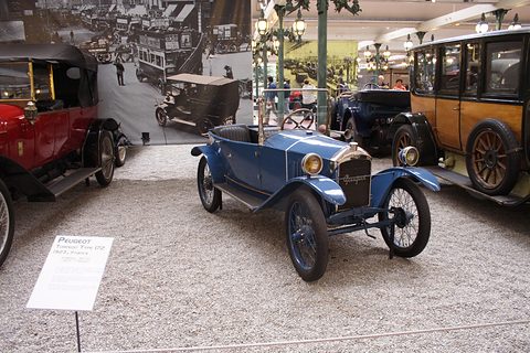 A small blue open car on gravel with a placard before it, a period street photograph on the wall behind. Opens a larger view