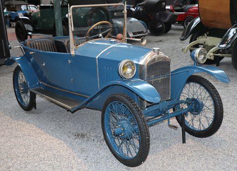 A blue open two-seater with a wooden-rimmed wheel and blue wire wheels on grey gravel. Opens a larger view