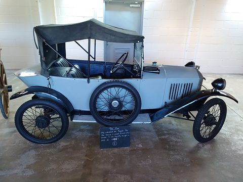 Side view of a pale grey two-seater with a black hood, louvred bonnet and wire wheels, a chalkboard sign in front. Opens a larger view