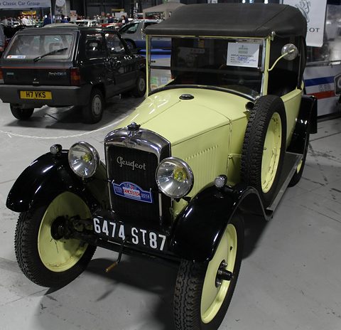A cream car with black wings and cream disc wheels, a Peugeot script radiator and a side spare wheel, a hood up. Opens a larger view