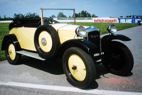 Side view of a cream open two-seater with black wings and cream disc wheels on tarmac, advertising boards behind. Opens a larger view