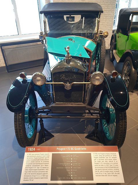 Front view of a turquoise tourer with a black hood, mascot and two lamps, a long museum placard in front. Opens a larger view