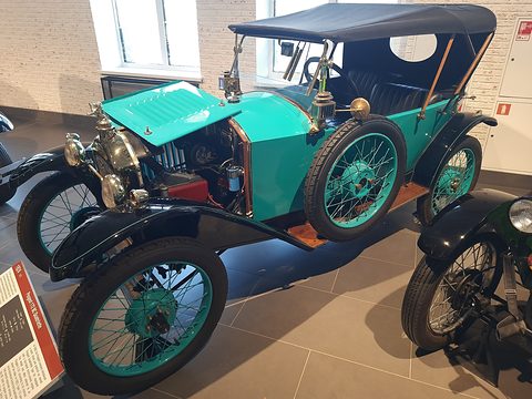 A turquoise tourer with a black hood and its bonnet raised, black wings and wire wheels on a tiled floor. Opens a larger view