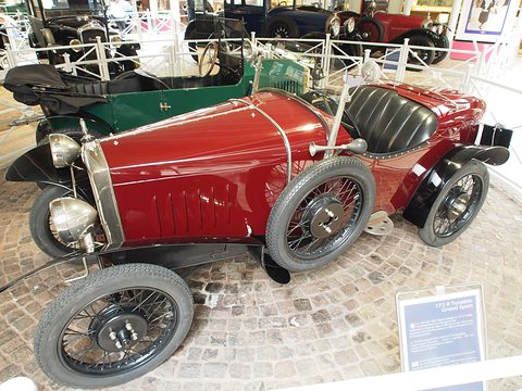 A dark red sports two-seater with a louvred bonnet, wire wheels and black seats on cobbles behind white railings. Opens a larger view