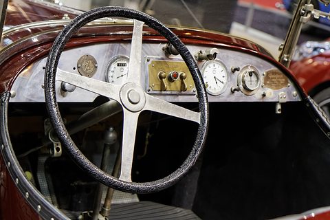A four-spoke steering wheel before an aluminium dashboard with round dials and a brass switch panel. Opens a larger view