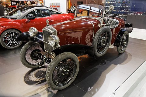 A dark red sports two-seater with a nickel radiator and black wire wheels on a grey stand, a modern red car behind. Opens a larger view