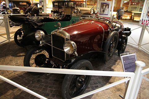 A maroon open car with a nickel radiator and black wings behind a white railing, a green car behind. Opens a larger view