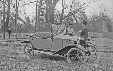 A small open two-seater with its windscreen up on muddy ground, two men in caps by the bonnet, bare trees behind.