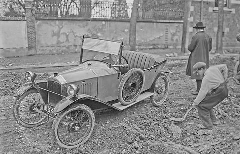 A narrow open two-seater with disc-like spoked wheels on churned ground, a man digging and another in a coat behind.