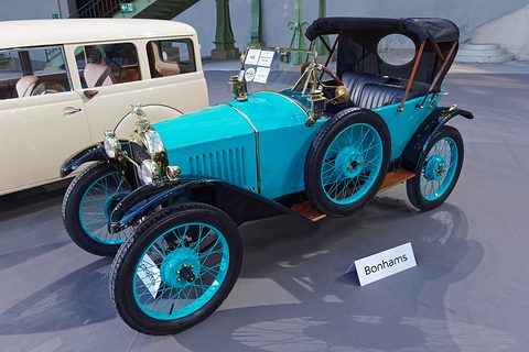 A bright turquoise open car with black wings, turquoise wire wheels and a black hood, a Bonhams sign on the floor. Opens a larger view