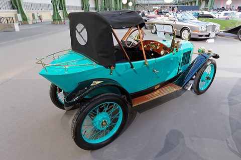 Rear quarter view of a turquoise car with a brass luggage rack, black hood and wooden steering wheel. Opens a larger view